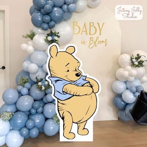 Puede incluir: Un recorte de Winnie the Pooh se encuentra frente a un telón de fondo con globos azules y blancos, vegetación y el texto "BABY in Bloom". La escena sugiere un tema de baby shower o guardería, con un ambiente alegre y festivo.