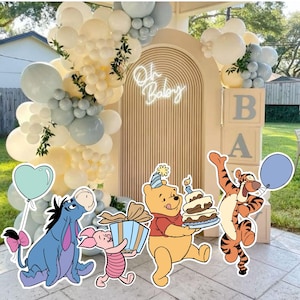 Puede incluir: Una decoración de baby shower con un fondo arqueado beige y un letrero de neón que dice Oh Baby, rodeado por una guirnalda de globos en crema, blanco y azul grisáceo. Incluye figuras de Ígor, Piglet, Winnie the Pooh con pastel y Tigger.