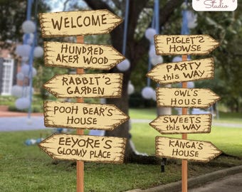 Op de afbeelding: Twee houten wegwijzers met meerdere richtingaanwijzers met teksten zoals Welcome, Hundred Acre Wood, Rabbit's Garden, Pooh Bear's House, Eeyore's Gloomy Place, Piglet's House, Party This Way, Owl's House, Sweets This Way en Kanga's House.