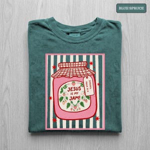 May include: A blue spruce t-shirt featuring a graphic of a pink jam jar with a red checkered lid. The jar is decorated with strawberry illustrations and the text "JESUS IS MY JAM!" with a small tag reading "SPREAD IT PSALM 34:8".