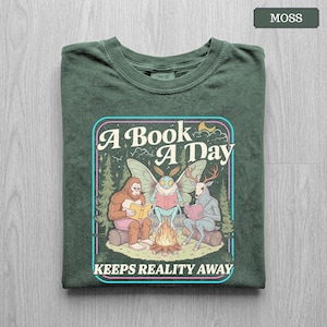 May include: A moss green t-shirt featuring a graphic print of a sasquatch, a moth, and a deer-like creature sitting around a campfire reading books. The text reads A Book A Day Keeps Reality Away, framed by a colorful border with trees and stars.
