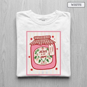 May include: A white t-shirt featuring a graphic of a pink strawberry jam jar with a red checkered lid. The label reads 'JESUS IS MY JAM!' and 'PSALM 34:8' on a small tag. The design includes red star accents and a light pink striped background.