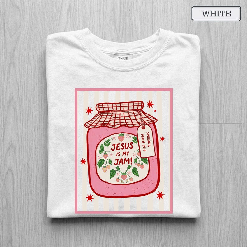 May include: A white t-shirt featuring a graphic of a pink strawberry jam jar with a red checkered lid. The label reads 'JESUS IS MY JAM!' and 'PSALM 34:8' on a small tag. The design includes red star accents and a light pink striped background.