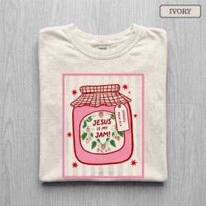 May include: An ivory t-shirt featuring a graphic of a pink strawberry jam jar with a red checkered lid. The label reads "JESUS IS MY JAM!" surrounded by strawberry illustrations. A small tag attached to the jar reads "SPREAD IT. PSALM 34:8".