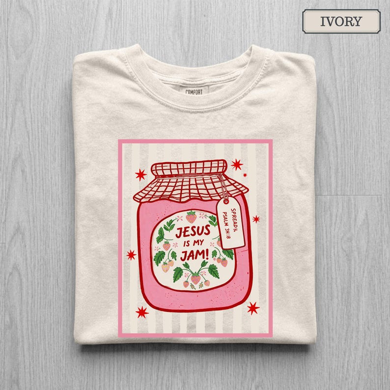 May include: An ivory t-shirt featuring a graphic of a pink strawberry jam jar with a red checkered lid. The label reads "JESUS IS MY JAM!" surrounded by strawberry illustrations. A small tag attached to the jar reads "SPREAD IT. PSALM 34:8".
