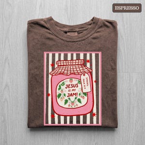 May include: An espresso brown t-shirt featuring a graphic of a pink strawberry jam jar on a striped background. The jar label reads "JESUS IS MY JAM!" with a small tag that says "SPREAD IT PSALM 34:8". The design includes red star accents.