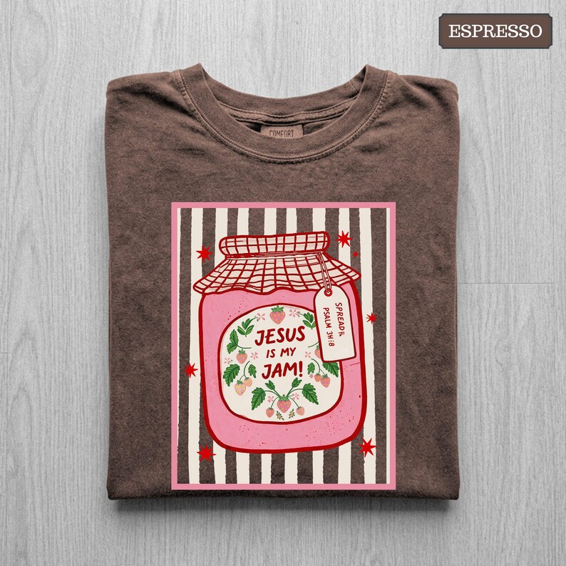 May include: An espresso brown t-shirt featuring a graphic of a pink strawberry jam jar on a striped background. The jar label reads "JESUS IS MY JAM!" with a small tag that says "SPREAD IT PSALM 34:8". The design includes red star accents.