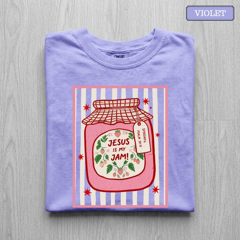 May include: A violet t-shirt featuring a graphic of a pink strawberry jam jar with a red checkered lid. The jar label reads "JESUS IS MY JAM!" with a small tag that says "SPREAD IT. PSALM 34:8". The background of the graphic has light blue stripes.