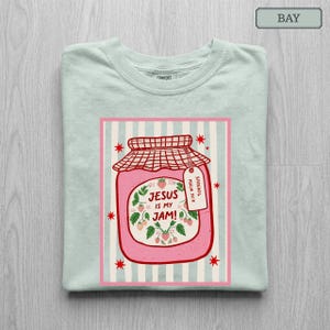 May include: A light green t-shirt featuring a graphic of a pink jar of strawberry jam with a red checkered lid. The jar label reads "JESUS IS MY JAM!" and includes a small tag that says "SPREAD IT PSALM 34:8". Red stars surround the jar.