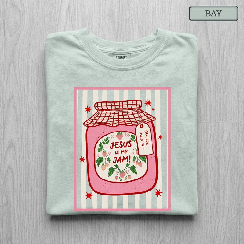 May include: A light green t-shirt featuring a graphic of a pink jar of strawberry jam with a red checkered lid. The jar label reads "JESUS IS MY JAM!" and includes a small tag that says "SPREAD IT PSALM 34:8". Red stars surround the jar.