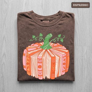 May include: A dark brown t-shirt featuring a graphic of a pumpkin constructed from stacked books. The pumpkin is orange and pink with a green stem and leaves. The text ESPRESSO appears in the top right corner of the image, indicating the color.