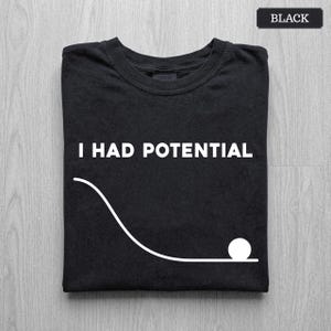 May include: A black crew neck t-shirt featuring the white text I HAD POTENTIAL centered above a white graphic of a curved line ending in a circle. The shirt is folded neatly and includes a small black label in the neck area with the word BLACK.