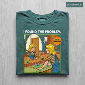 May include: A dark teal t-shirt featuring a graphic of a person with blonde hair wearing a striped shirt and blue overalls, standing at a bathroom sink. The text reads I FOUND THE PROBLEM and Inner Reflection in a comic book art style.