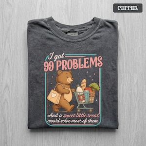 May include: A charcoal gray t-shirt featuring a graphic of a brown bear carrying a shopping bag and pushing a cart filled with groceries. The text reads, I got 99 PROBLEMS And a sweet little treat would solve most of them, in pink and teal.