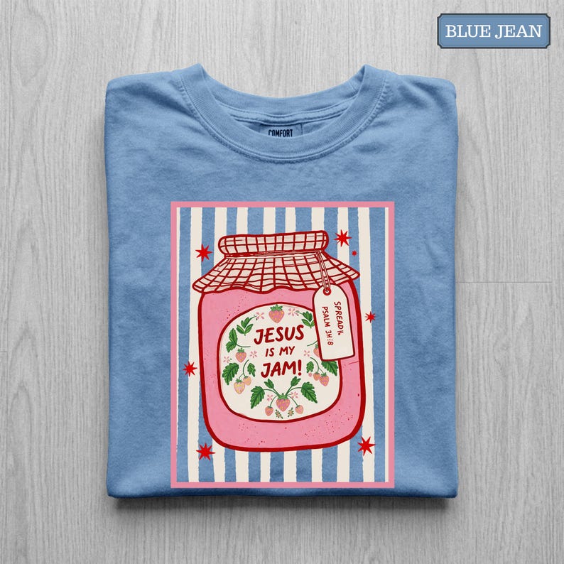 May include: A blue jean colored t-shirt featuring a graphic of a pink jam jar with a red checkered lid. The jar label reads "JESUS IS MY JAM!" surrounded by strawberry illustrations and a small tag that says "SPREAD IT. PSALM 34:8".