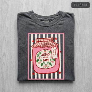 May include: A charcoal gray t-shirt featuring a graphic of a pink strawberry jam jar with a red checkered lid. The jar label reads "JESUS IS MY JAM!" and "SPREAD IT PSALM 34:8" with strawberry illustrations. The background has vertical black stripes.