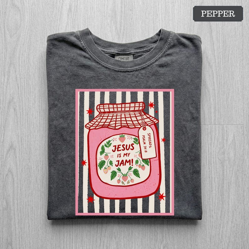May include: A charcoal gray t-shirt featuring a graphic of a pink strawberry jam jar with a red checkered lid. The jar label reads "JESUS IS MY JAM!" and "SPREAD IT PSALM 34:8" with strawberry illustrations. The background has vertical black stripes.