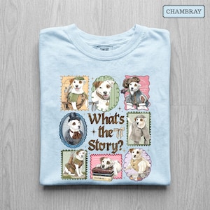 May include: A light blue t-shirt featuring a graphic print of six Jack Russell terriers dressed in historical costumes within decorative frames. The central text reads What's the Story? with small star and ribbon accents surrounding the dog illustrations.