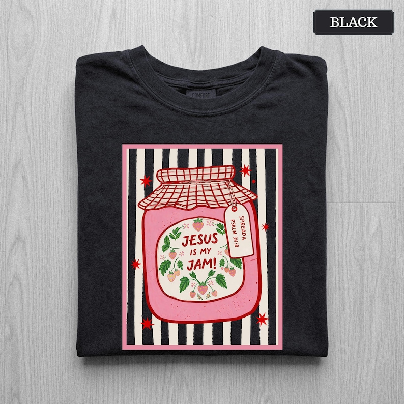May include: A black t-shirt featuring a graphic of a pink strawberry jam jar on a black and white striped background. The jar label reads "JESUS IS MY JAM!" with a small tag that says "SPREAD IT. PSALM 34:8". The design includes red stars.