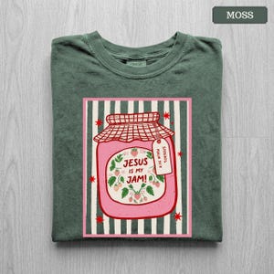 May include: A moss green t-shirt featuring a graphic of a pink strawberry jam jar. The jar has a red checkered lid, a tag reading "Spread it Psalm 34:8", and the text "Jesus is my jam!" surrounded by strawberry vines on a striped background.