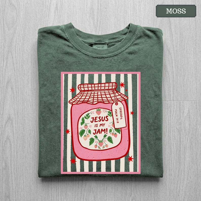 May include: A moss green t-shirt featuring a graphic of a pink strawberry jam jar. The jar has a red checkered lid, a tag reading "Spread it Psalm 34:8", and the text "Jesus is my jam!" surrounded by strawberry vines on a striped background.