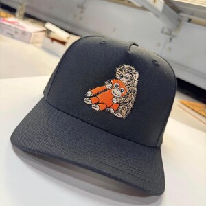 K&ouml;nnte beinhalten: Schwarze Baseballkappe mit einem gestickten Design von zwei Cartoon-Affen, die sich umarmen. Die Kappe hat einen gebogenen Schirm und eine strukturierte Krone. Die Affen sind orange und braun, die Kappe selbst ist einfarbig schwarz.