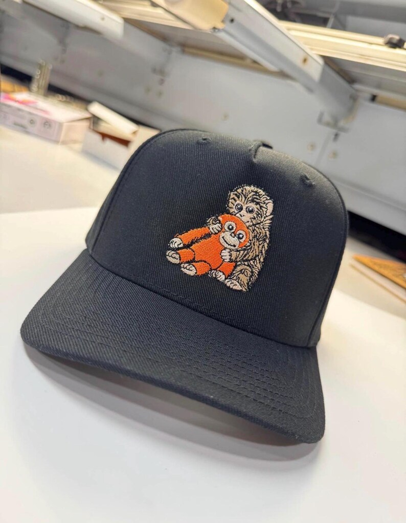 K&ouml;nnte beinhalten: Schwarze Baseballkappe mit einem gestickten Design von zwei Cartoon-Affen, die sich umarmen. Die Kappe hat einen gebogenen Schirm und eine strukturierte Krone. Die Affen sind orange und braun, die Kappe selbst ist einfarbig schwarz.