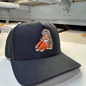 K&ouml;nnte beinhalten: Schwarze Baseballkappe mit gebogenem Schirm. Die Kappe hat ein gesticktes Design von zwei Zeichentieren, einem orangen Affen und einem braun-wei&szlig;en Tier. Die Kappe ist aus strapazierf&auml;higem Stoff gefertigt und hat eine strukturierte Passform.