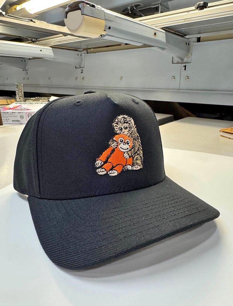 K&ouml;nnte beinhalten: Schwarze Baseballkappe mit gebogenem Schirm. Die Kappe hat ein gesticktes Design von zwei Zeichentieren, einem orangen Affen und einem braun-wei&szlig;en Tier. Die Kappe ist aus strapazierf&auml;higem Stoff gefertigt und hat eine strukturierte Passform.