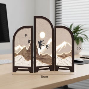 Könnte beinhalten: Ein dekorativer dreiteiliger Bildschirm mit einem von der Natur inspirierten Design. Der Bildschirm hat einen braunen Rahmen und eine Landschaftsszene mit Bergen, Vögeln und einer Sonne. Der Bildschirm ist 40 cm breit und 30 cm hoch.