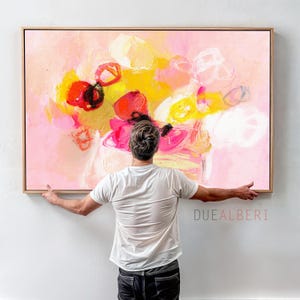 Puede incluir: Un gran cuadro rectangular sobre lienzo con pinceladas abstractas en tonos rosa, amarillo, rojo y blanco, enmarcado en madera clara. La obra muestra texturas superpuestas y marcas circulares. El texto DUEALBERI está impreso en la parte inferior derecha.