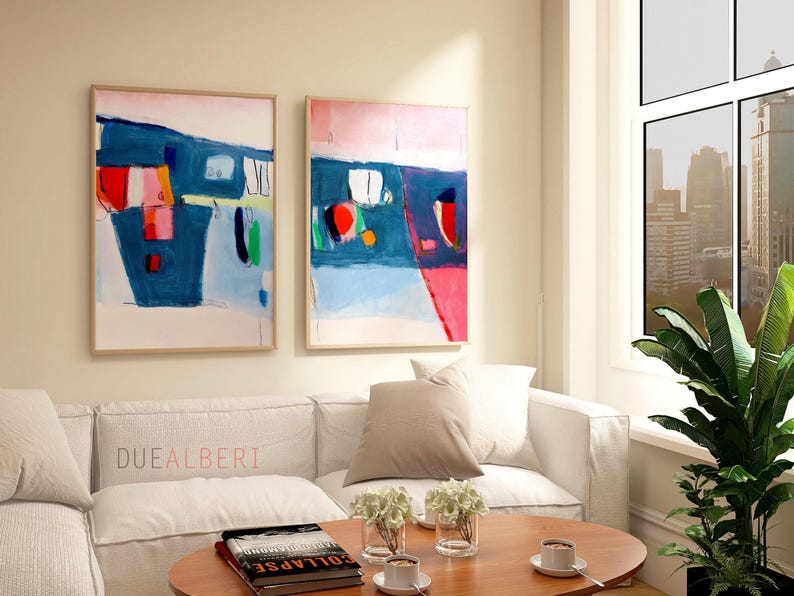 May include: Two framed abstract paintings with blue, red, and white shapes hang above a white sofa. A round wooden coffee table holds books, cups, and flowers. The text "DUEALBERTI" is on the sofa.