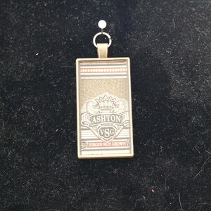 May include: A rectangular pendant with a gold-colored frame. The pendant features the text "ASHTON VSG" in a shield design, with a crown above the text. The background is a mix of gold and red stripes. A small metal loop is at the top.