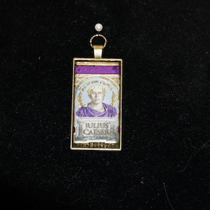 May include: A rectangular pendant with a gold-colored frame. The pendant features a portrait of Julius Caesar, with the text "JULIUS CAESER" below the portrait. The top of the pendant has a purple section. The pendant is suspended from a gold-colored chain.