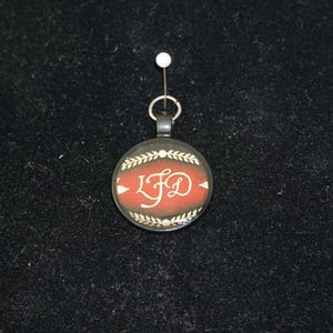 May include: A round pendant with a red center and the letters "LFD" in white script. The pendant is encircled by a laurel wreath design and has a silver-colored bail and chain. The pendant is suspended from a white pin.