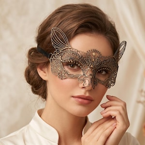 May include: A metallic gold filigree masquerade mask featuring intricate scrollwork patterns and pointed cat-ear silhouettes. The mask covers the eye area and is secured with a thin black ribbon strap, showcasing a detailed openwork design.