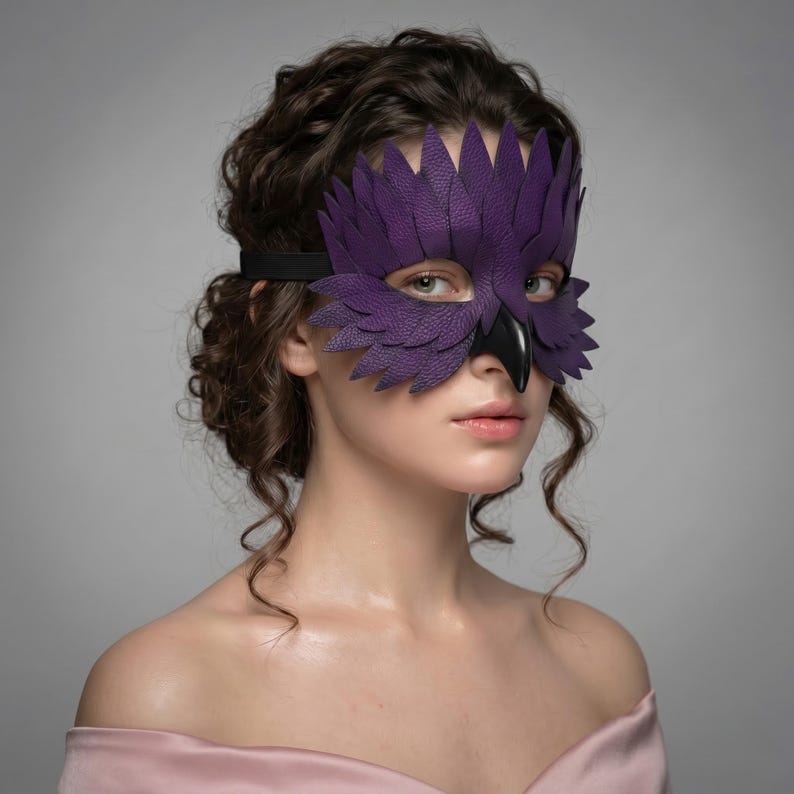 May include: A purple leather owl mask with feather detailing and a black beak, secured with a black elastic band. The mask is worn by a person with curly brown hair. The mask is designed to cover the eyes and nose.