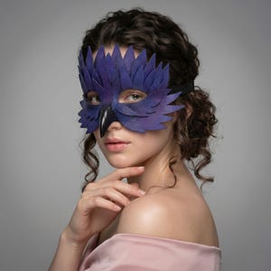 May include: A purple leather mask with a bird-like design, featuring layered feather details and a black beak. The mask is secured with a black elastic band. The mask is a costume accessory.