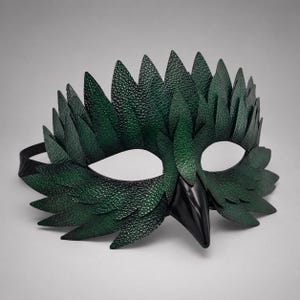 May include: A handcrafted green leather mask with a bird-like design. The mask features a textured surface, pointed leaf-like shapes, and a black beak. The eye openings are white, and a black strap secures the mask.
