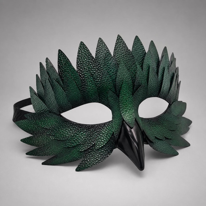 May include: A handcrafted green leather mask with a bird-like design. The mask features a textured surface, pointed leaf-like shapes, and a black beak. The eye openings are white, and a black strap secures the mask.