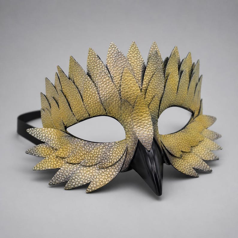 May include: A handcrafted bird mask with a textured, gold and grey feather design. The mask features a black beak and eye openings, attached to a black elastic band. The mask is designed for costume or theatrical use.