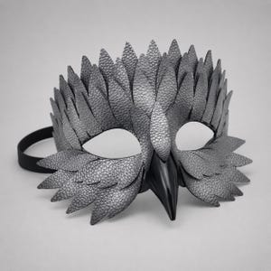 May include: A silver-grey textured mask with a bird-like design. The mask has a prominent beak and feather-like details around the eyes and forehead. A black elastic band is attached. Suitable for costume parties or theatrical performances.
