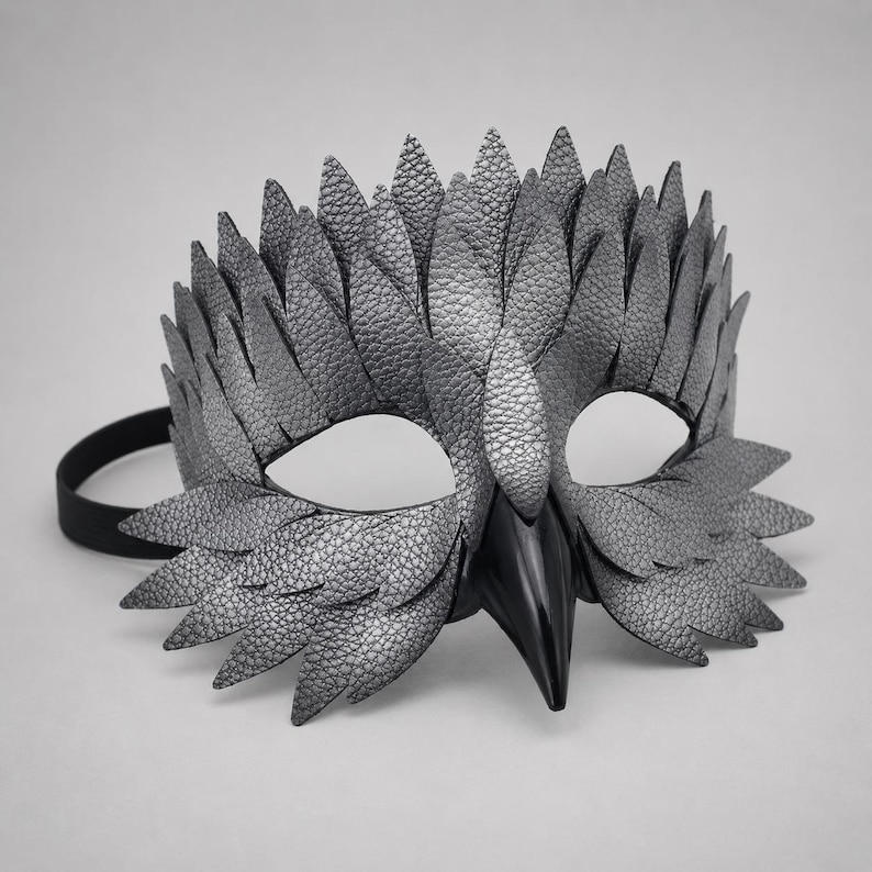 May include: A silver-grey textured mask with a bird-like design. The mask has a prominent beak and feather-like details around the eyes and forehead. A black elastic band is attached. Suitable for costume parties or theatrical performances.