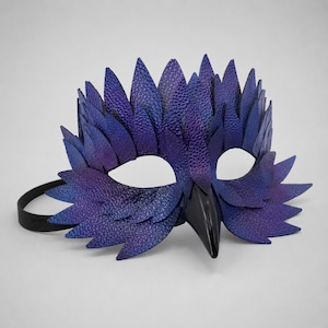 May include: A handcrafted, purple and blue leather mask with a bird-like design. The mask features a textured finish, pointed feather-like details, and a black beak. It has a black elastic strap for wearing.