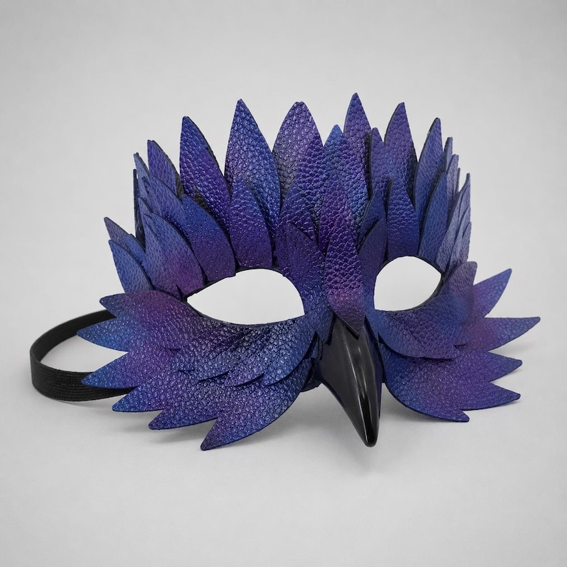May include: A handcrafted, purple and blue leather mask with a bird-like design. The mask features a textured finish, pointed feather-like details, and a black beak. It has a black elastic strap for wearing.