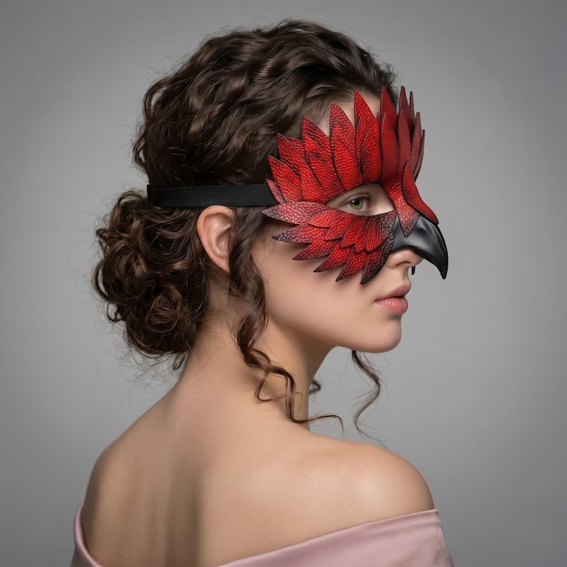May include: A red and black leather mask shaped like a bird's head. The mask has a detailed feather design in red and a black beak. It is held in place with a black elastic band.
