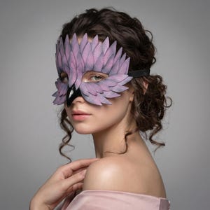 May include: A lavender and black bird-shaped mask with feather detailing. The mask has a black elastic band and a glossy black beak. The mask is worn over the eyes and nose. The mask is made of leather.