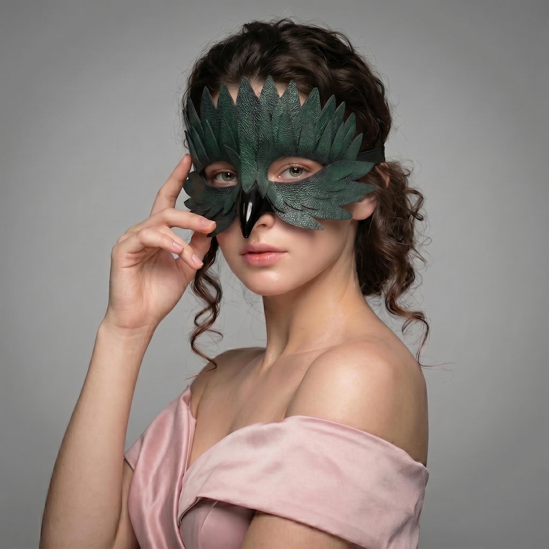 May include: A dark green leather mask with a bird-like design. The mask features a beak and feather-like details, with a black strap. The mask is worn by a person with curly brown hair and a pink dress.