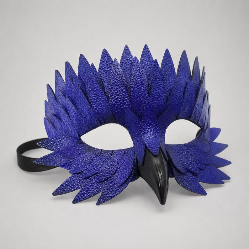 May include: A dramatic blue and black masquerade mask. The mask has a textured, feather-like design in blue, with a black beak and eye openings. A black elastic band secures the mask.