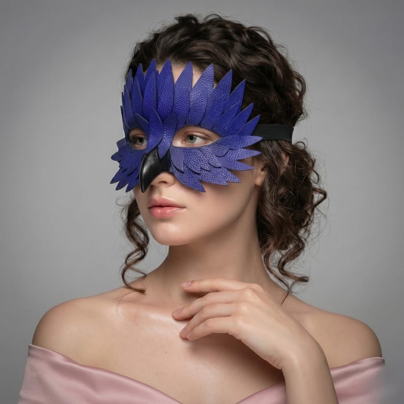 May include: A blue leather bird mask with a black beak and elastic strap. The mask, with a textured finish, is worn by a person with curly brown hair. The mask is designed to cover the eyes.